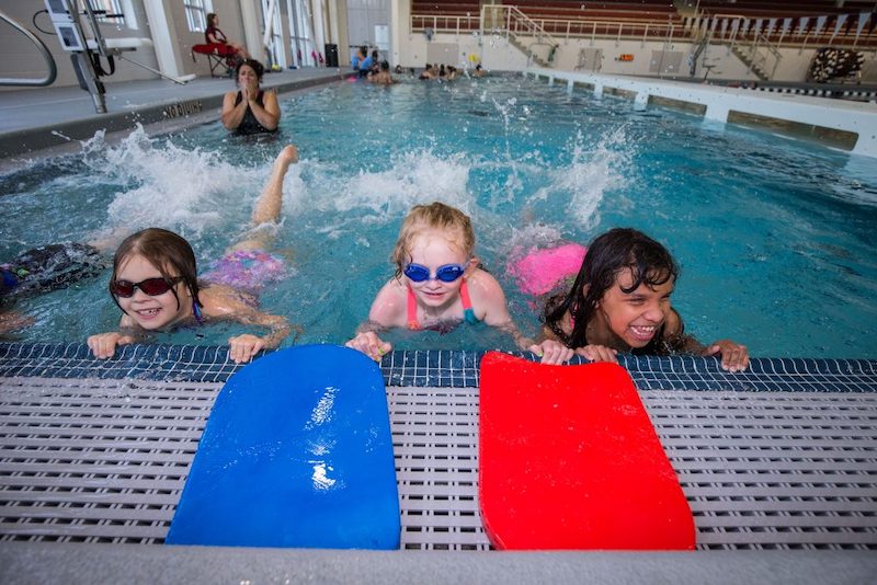 Three young girls holding on to the edge of a pool kicking their legs behind them.