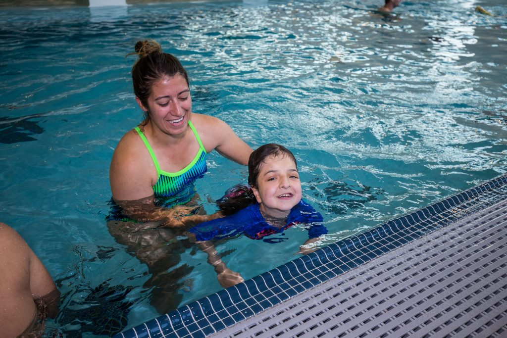 Young girl holding onto the edge of the pool with a female volunteer behind her.