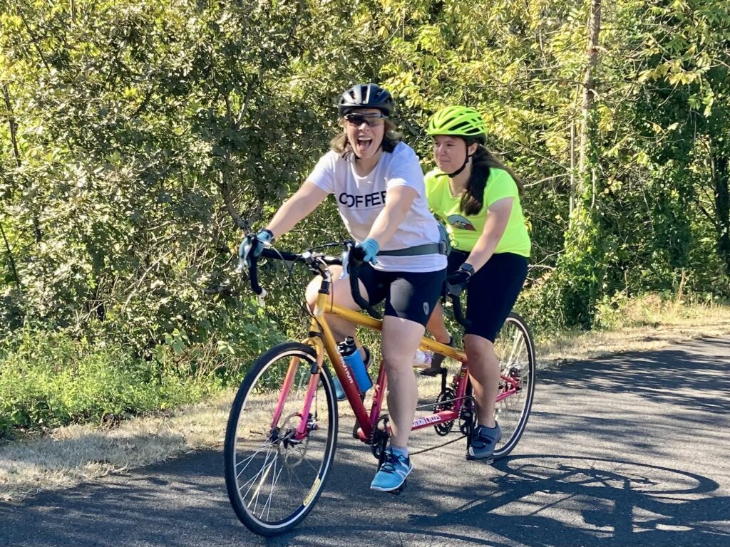Female athlete and guide riding tandem on a tree lined path.